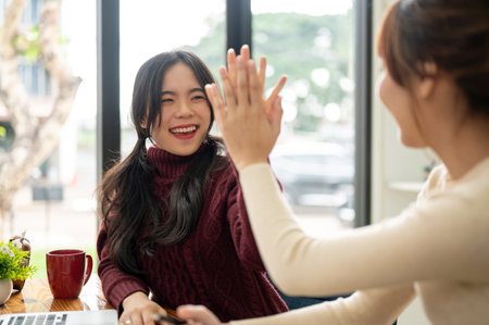 A cheerful young Asian woman is giving a high five to her friend to cheer her up and celebrate...