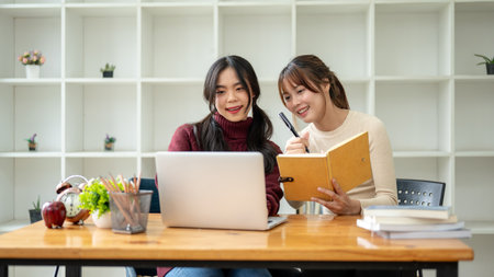 Two cheerful young Asian female college students are doing a homework and working on a co-project at...