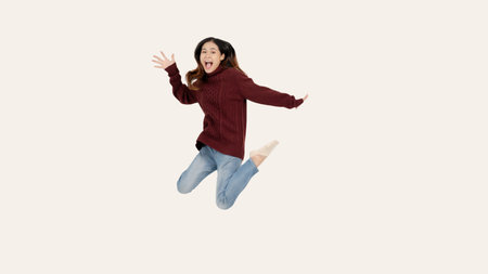 An overjoyed and pretty Asian woman in a red sweater is jumping with joy over an isolated white...
