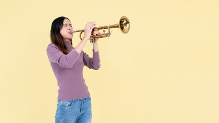 A beautiful and talented Asian woman is playing the trumpet while standing against an isolated yellow studio background. musician, music, instrumentの写真素材