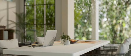 A laptop computer on a dining table in a modern dining room with a beautiful green nature view...