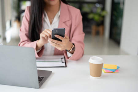 Cropped image of a beautiful and professional Asian businesswoman in a pink business suit using her smartphone at her desk in the office.の写真素材