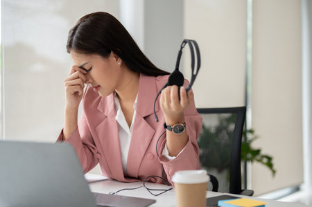 A stressed Asian female call centre agent or customer service operator sits at her desk, feeling tired and overwhelmed.の写真素材