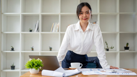 An attractive and confident Asian businesswoman or female manager smiling at the camera while standing at her desk in her private office.の写真素材