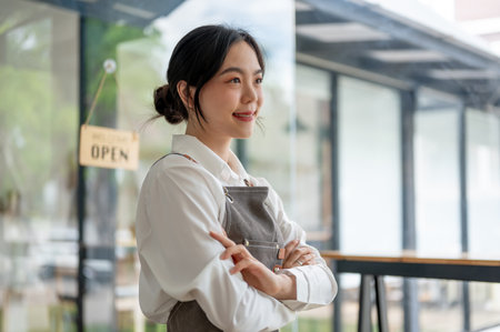 A successful and confident Asian female small business owner in an apron stands with her arms crossed in her cafe. SME, small business, worker, hospitality serviceの写真素材