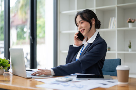 A confident and beautiful Asian businesswoman or female accountant is talking on the phone with her client while working at her desk in the office.の写真素材