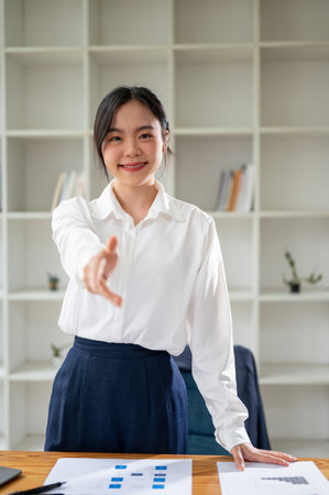 A portrait of an attractive and confident Asian businesswoman or female executive manager stands at her desk with a handshake gesture. Handshake, welcome, meetingの写真素材
