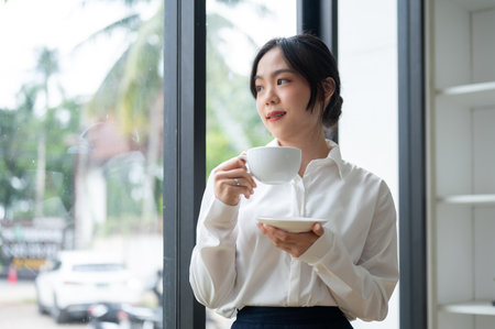 A beautiful and relaxed Asian businesswoman or female executive manager sipping coffee while standing by the window in her office.の写真素材