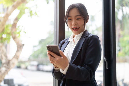 A beautiful and cheerful Asian businesswoman in a formal business suit using her smartphone by the window in her office, receiving good news, unexpected SMS or emailsの写真素材