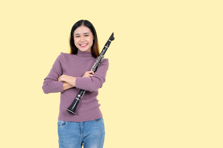 An attractive and joyful Asian woman holding a clarinet, stands against an isolated yellow background. Musician, musical student, leisure, and hobbyの写真素材