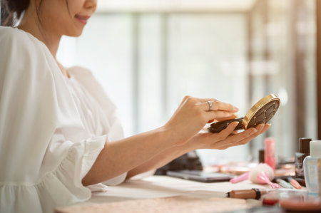 Close-up image of a beautiful Asian woman holding a compact powder or cushion, using pressed powder, doing make-up at home. rear viewの写真素材
