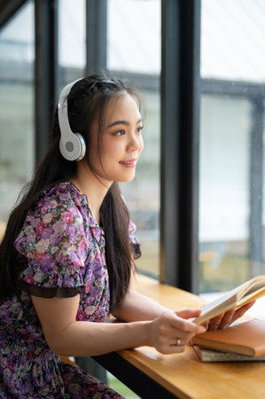 A portrait of a beautiful and charming young Asian woman in a cute dress listening to music and reading a book while relaxing in a coffee shop.の写真素材