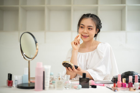 A beautiful and charming young Asian woman applies cushion or press powder on her face, getting ready at her vanity dress in her room. Lifestyle conceptの写真素材