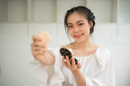 An attractive Asian female beauty influencer applying her makeup, showing her new cosmetic product to a camera and creating her makeup tutorial video at home.の写真素材