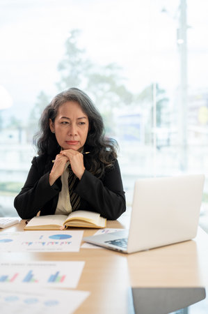 A portrait of a professional and serious senior Asian businesswoman looking at her laptop screen...