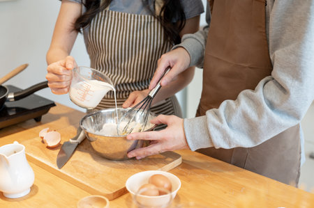 Close-up image of a couple mixing pancake flour and pouring milk into a mixing bowl while making...