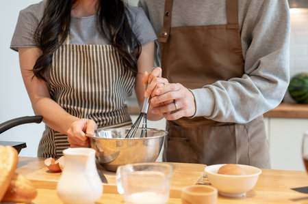 Close-up image of a lovely and happy young Asian couple in an apron is whisking egg and flour together, having a cooking date at home, and enjoying making pancakes in the kitchen.の写真素材