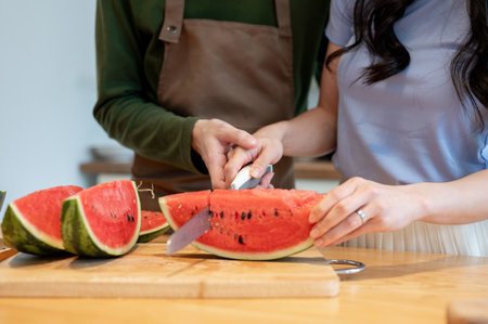 Close-up image of a boyfriend helping and teaching his girlfriend to cut a watermelon while cooking in the kitchen together.の写真素材