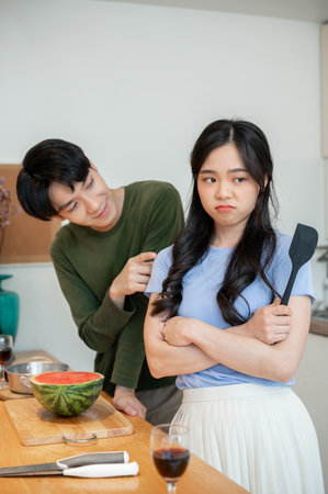 A cute, clingy young Asian boyfriend is asking for forgiveness from his angry girlfriend after arguing while cooking in the kitchen together.の写真素材