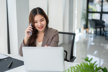 A cheerful and beautiful Asian businesswoman or female sales agent is looking at her laptop screen while talking on the phone and providing a positive experience for her client.