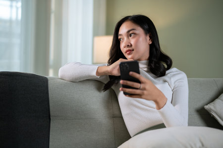 A gorgeous, thoughtful Asian woman is looking out the window while sitting on a sofa in the living room with her smartphone in her hand, thinking or planning something in her mind.の写真素材