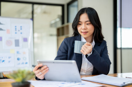 A beautiful and professional Asian businesswoman or female accountant is working at her desk in a modern office, using a tablet while sipping her morning coffee.の写真素材
