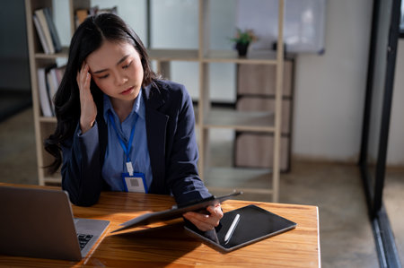 A thoughtful and focused Asian businesswoman or female manager in a formal business suit is feeling tired from overwork while reading documents at her desk in the office.の写真素材