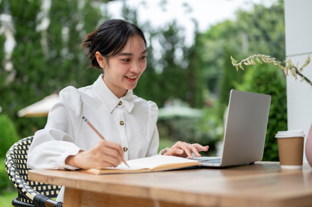 A beautiful Asian woman using her laptop and taking notes on her notebook while sitting at a table in a beautiful garden. college student, freelancer, working remotelyの写真素材