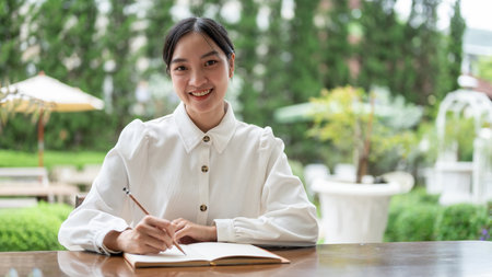 Portrait of a beautiful young Asian woman is smiling at the camera while sitting at a table with her diary in a beautiful green garden.の写真素材