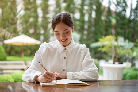 A beautiful and happy young Asian woman is keeping her diary, listing her ideas, or writing something in her book while sitting at a table in her beautiful green backyard. Lifestyle conceptの写真素材