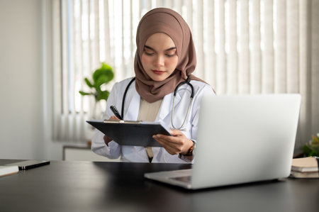 A professional and beautiful Asian Muslim female doctor in a uniform and hijab is focusing on checking her medical case information on a clipboard, working at her desk in an examination room.の写真素材