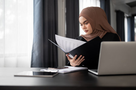 A professional adult Asian Muslim businesswoman or female manager focuses on examining business reports in a binder, working at her desk in her private office.の写真素材