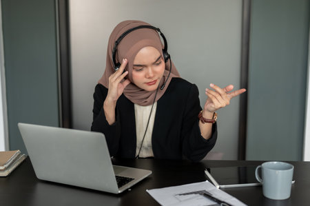 A confused and thoughtful Asian Muslim female customer call centre operator wearing a headset and hijab resolves customer issues over the call, consulting with a client over the phone.の写真素材