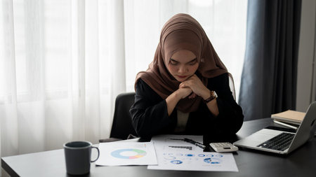 A focused and thoughtful millennial Asian Muslim businesswoman wearing a hijab is examining and working on business reports at her desk in the office.の写真素材