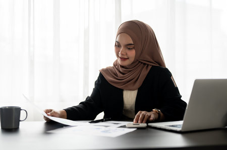 A professional Asian Muslim businesswoman or female manager in a hijab is working on financial reports, examining data on reports at her desk in a modern office.の写真素材