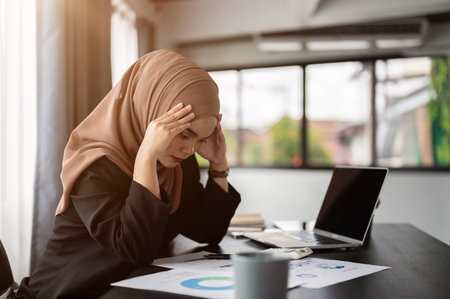 A stressed and thoughtful millennial Asian Muslim businesswoman in a hijab is reading and examining business reports with a serious face, worried about her project, working at her desk in the office.の写真素材