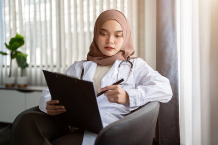 A focused and professional Asian Muslim female doctor in a uniform and hijab is checking her medical cases on a medical clipboard while sitting in her office at a hospital.の写真素材
