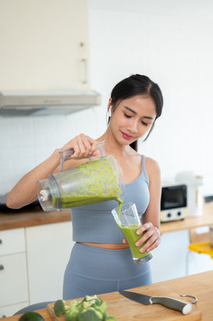 A portrait of a beautiful and happy Asian woman in gym clothes is pouring her healthy green smoothie into a glass, making her healthy breakfast in the kitchen after a workout.の写真素材