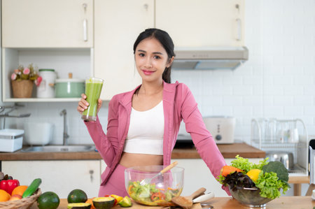 A gorgeous and slim Asian woman in gym clothes is holding a glass of a healthy green smoothie while standing at a kitchen table with fresh fruits and vegetables on the table. Healthy lifestyle conceptの写真素材