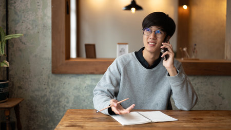 A happy young Asian man is talking on the phone with someone while working remotely at a coffee shop...
