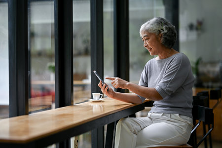 A relaxed and happy Asian retired lady in casual clothes enjoys the app on her smartphone while sitting in a coffee shop. Retirement lifestyle conceptの写真素材