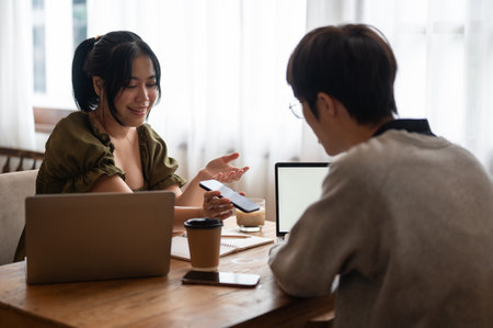 A charming Asian female college student is talking and showing something on her phone to a friend while sitting in a coffee shop together.の写真素材