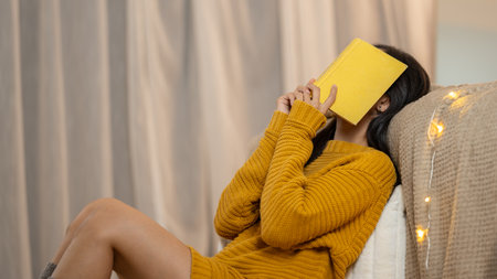 Side view image of a tired Asian woman in a cosy knitted sweater covering her face with a book, feeling sleepy or tired while reading a book in her bedroom.の写真素材