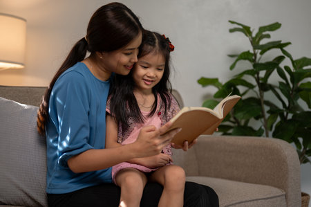 A caring young Asian mom is embracing her daughter while reading a book, telling a story, or reading a fairy tale on a couch in the living room together.の写真素材