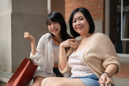 Two beautiful and happy young Asian women are smiling at the camera, sitting on the stairs with their shopping bags, and enjoying their shopping day in the city.の写真素材