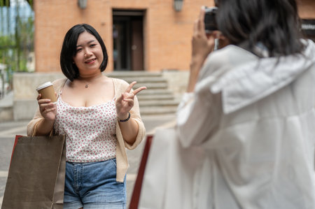 A happy Asian plus-size woman is being taken a photo by her friend while enjoying shopping day in the city together, carrying shopping bags, showing the peace hand sign and smiling at the camera.の写真素材