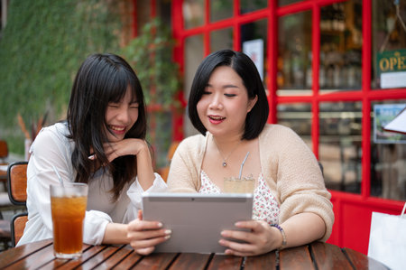 Two happy young Asian female friends are watching a video on a tablet together while relaxing at an outdoor table of a coffee shop in the city.の写真素材