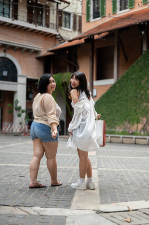 Back view image of two happy Asian female friends are holding their shopping bags, enjoying walking and shopping in the city street together on the holiday.の写真素材