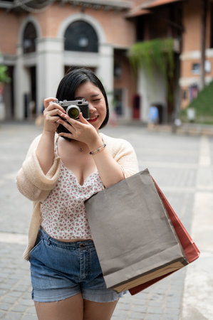 A beautiful and happy Asian plus-size woman is carrying her shopping bags and using her film camera to take a photo while enjoying her shopping day in the city.の写真素材
