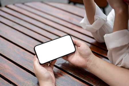 Close-up image of a woman holding a white-screen smartphone mockup in a horizontal position while sitting at a table with her friend. watching video, playing game, using mobile appの写真素材